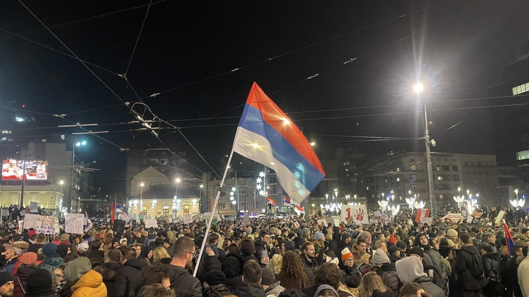 Folla di manifestanti di notte in piazza Slavija a Belgrado, con una bandiera serba in primo piano e cartelli di protesta