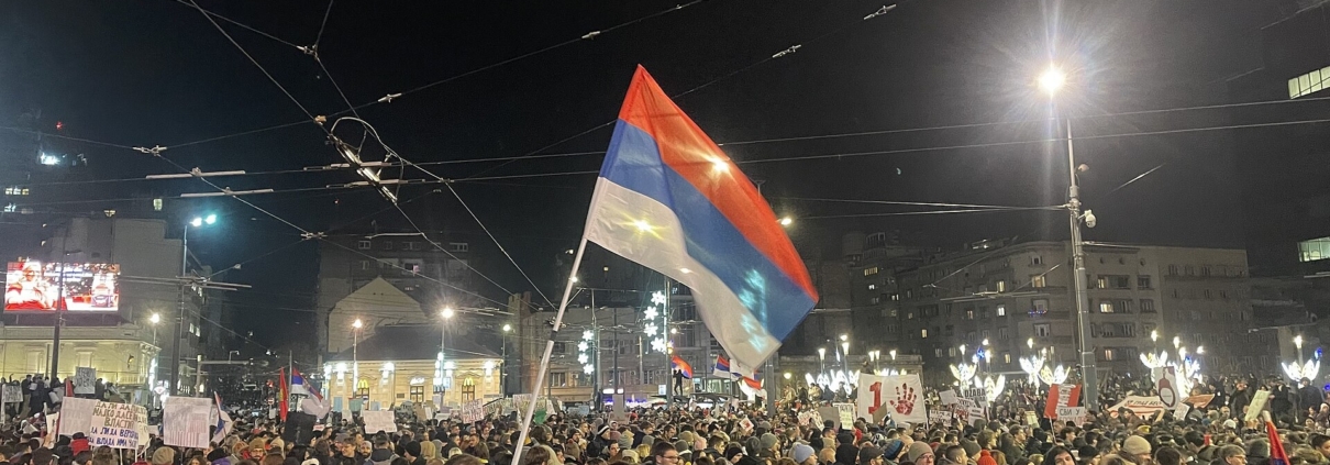 Folla di manifestanti di notte in piazza Slavija a Belgrado, con una bandiera serba in primo piano e cartelli di protesta