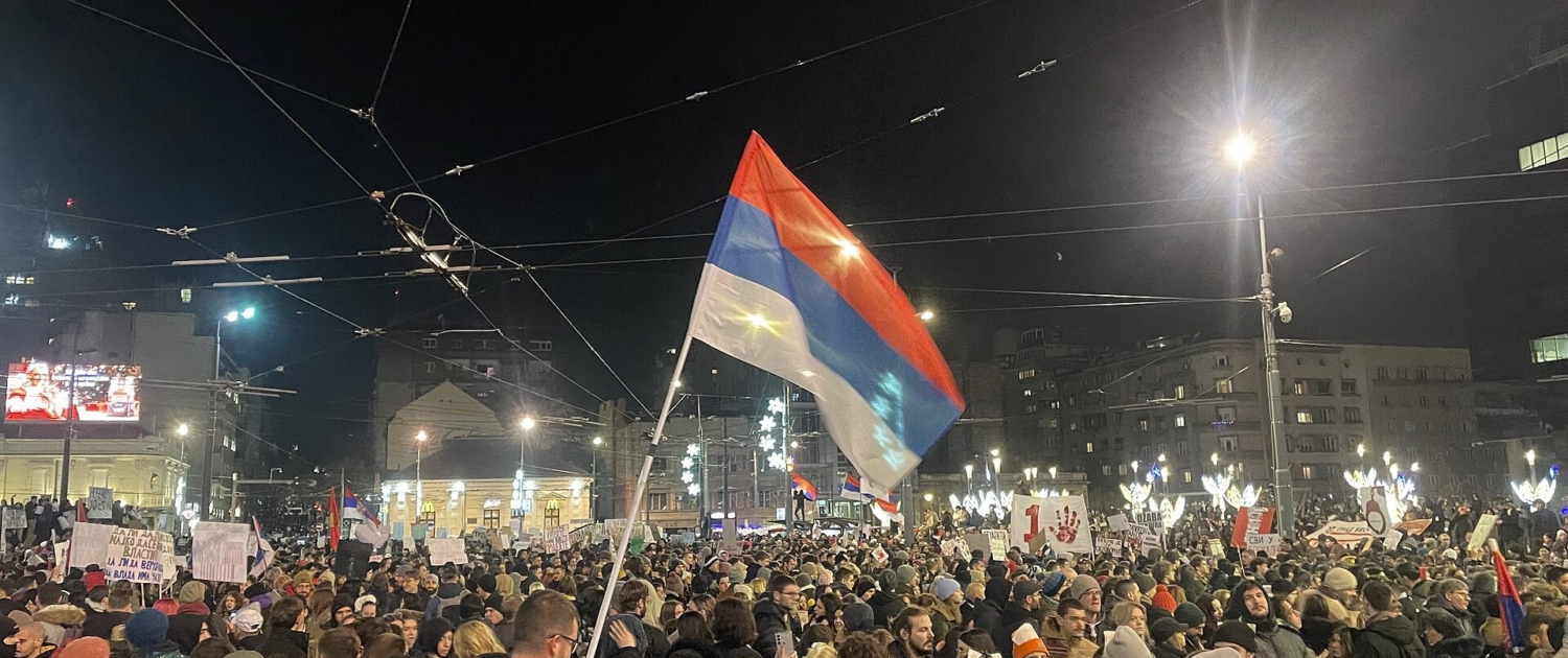 Folla di manifestanti di notte in piazza Slavija a Belgrado, con una bandiera serba in primo piano e cartelli di protesta