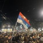 Folla di manifestanti di notte in piazza Slavija a Belgrado, con una bandiera serba in primo piano e cartelli di protesta