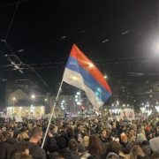 Belgrado, piazza Slavija, 22 dicembre 2024 Folla di manifestanti di notte in piazza Slavija a Belgrado, con una bandiera serba in primo piano e cartelli di protesta