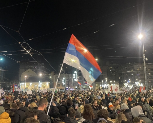 Folla di manifestanti di notte in piazza Slavija a Belgrado, con una bandiera serba in primo piano e cartelli di protesta